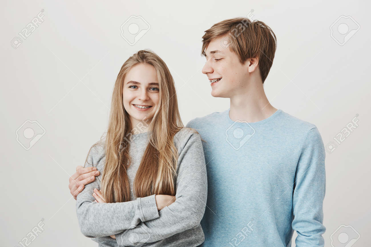 고운 머리를 가진 키 큰 매력적인 젊은 남자, 포옹하고 여자 친구를 바라보고, 그가 그녀를 사랑하는 방법을 생각하면서 활짝 웃고, 회색  벽 위에 서 있습니다. 로열티 무료 사진, 그림, 이미지 그리고 스톡포토그래피. Image