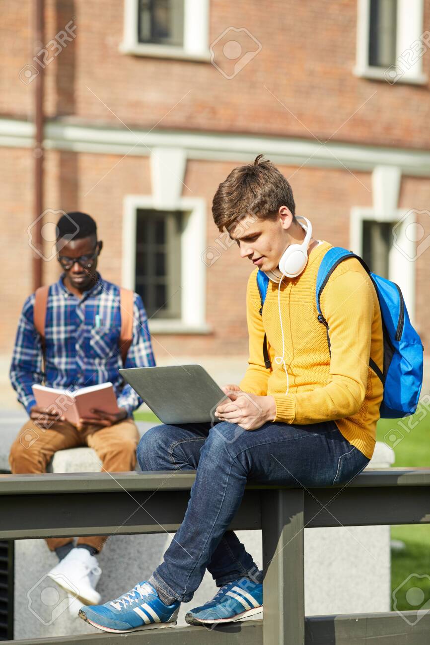 Full length portrait of contemporary college student using laptop sitting on table outdoors in campus, copy space - 128446461 Full length portrait of contemporary college student using laptop sitting on table outdoors in campus, copy space - 128446461