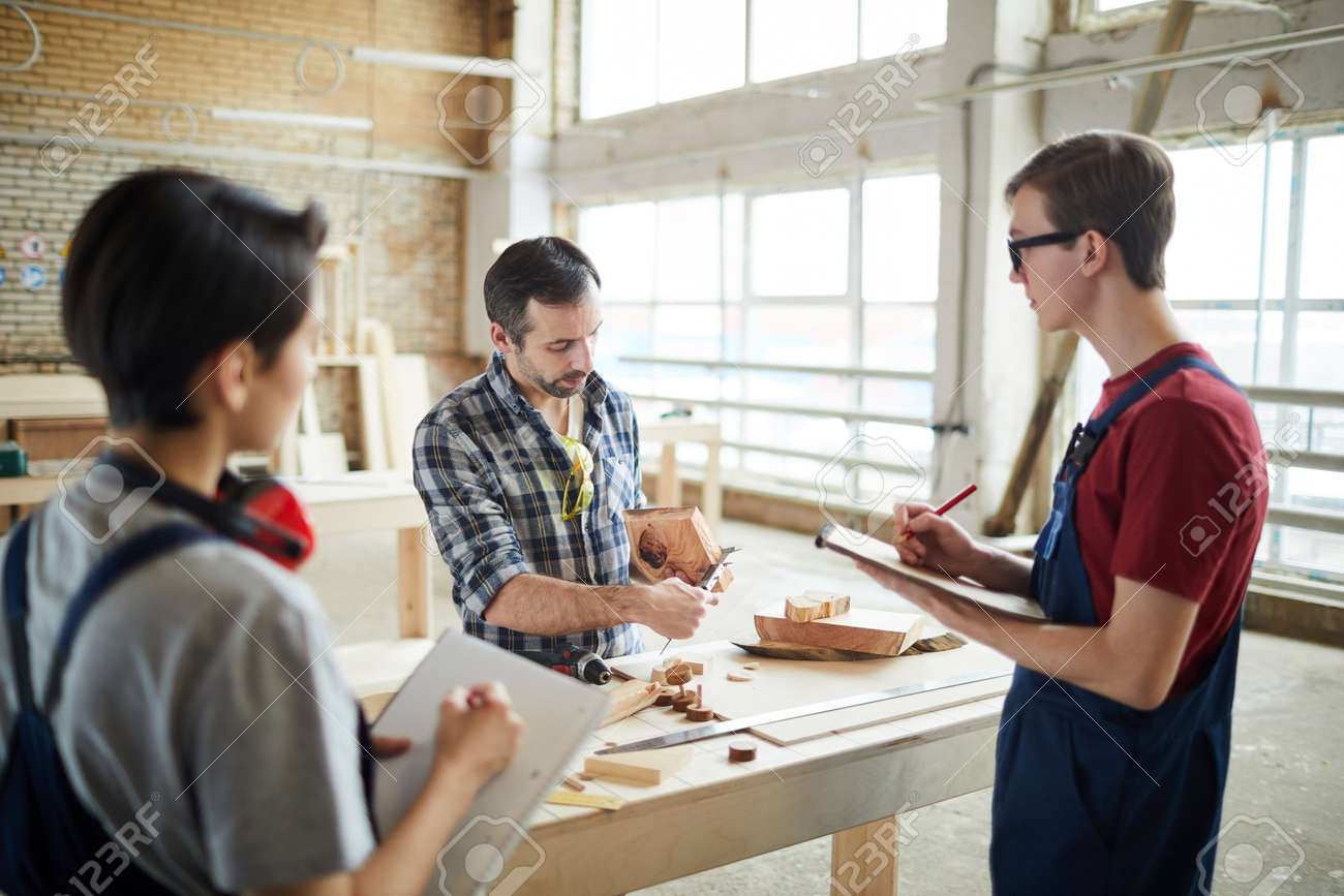 Waist up portrait of mature carpenter teaching young apprentices in workshop - 129172607 Waist up portrait of mature carpenter teaching young apprentices in workshop - 129172607