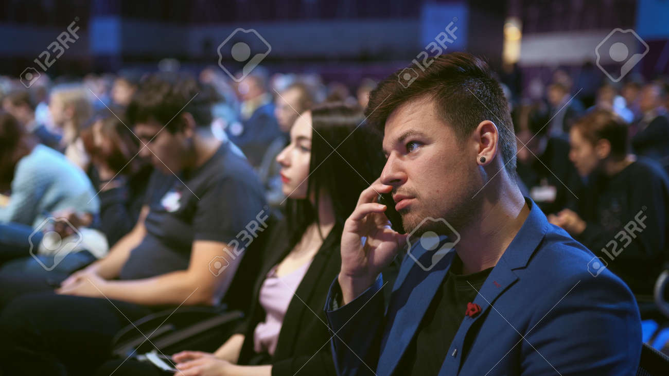 Business consultant talk on phone in crowd audience. Person negotiating on mobile in auditorium. Business man talking on phone economic forum. People discusses strategy partner in viewer audience. - 156059473 Business consultant talk on phone in crowd audience. Person negotiating on mobile in auditorium. Business man talking on phone economic forum. People discusses strategy partner in viewer audience. - 156059473