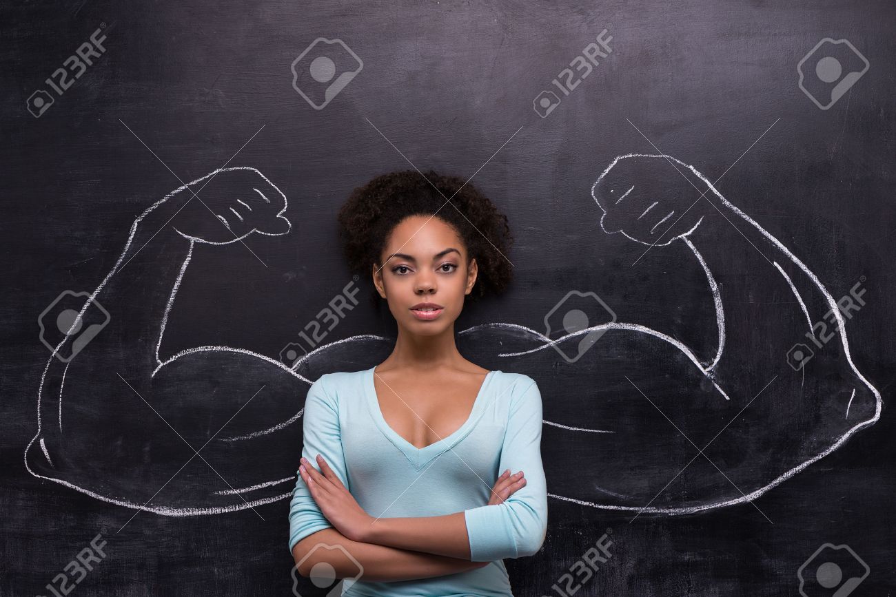 真剣にカメラを見て黒板背景に若いアフリカ系アメリカ人女性の面白い画像 黒板に描かれた 2 つの強力な筋肉の腕 の写真素材 画像素材 Image