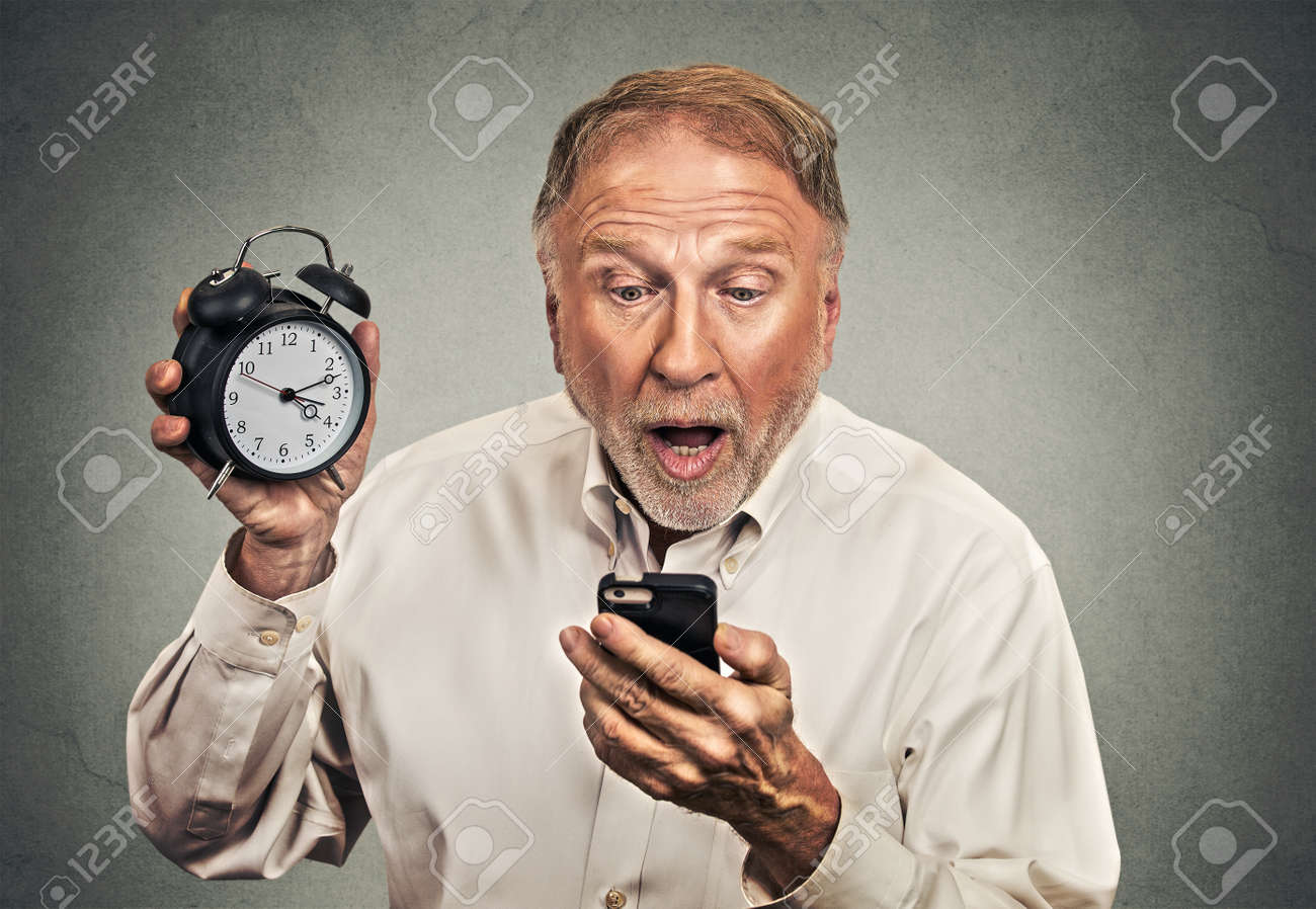 肖像画は 分離会議灰色の壁背景に遅刻面白い表情でスマート フォンを見ての目覚まし時計で商売していた男を驚かせた 人物顔表現 感情 感情 の写真素材 画像素材 Image