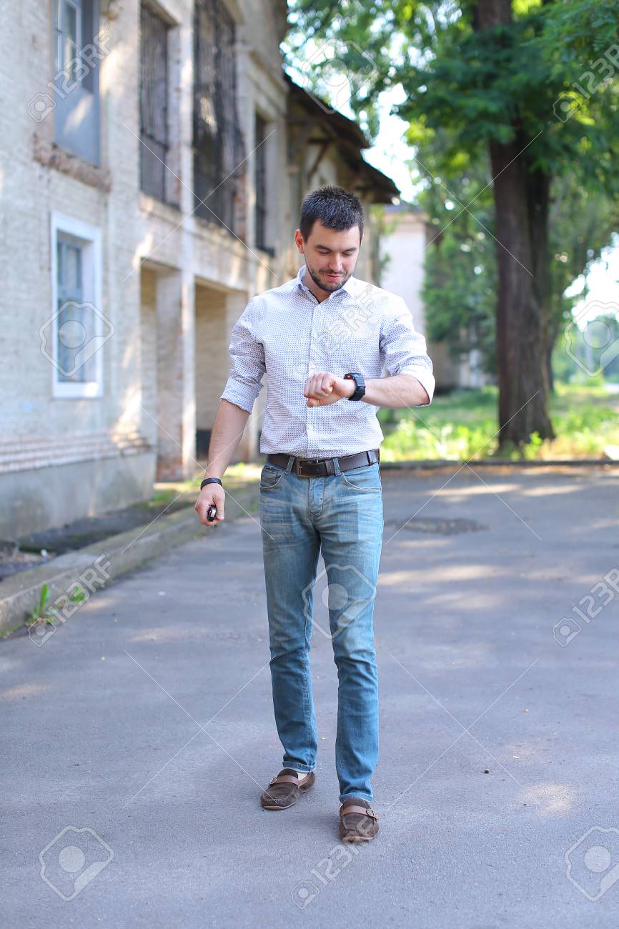 Hombres Jóvenes Con Barba Hombre Tipo Va, Caminando A Lo Largo De Carreteras Y Mirando El Reloj En Brazo. Joven Vestido De Camisa Blanca Con Franja Roja, Pantalones Vaqueros Y Mocasines
