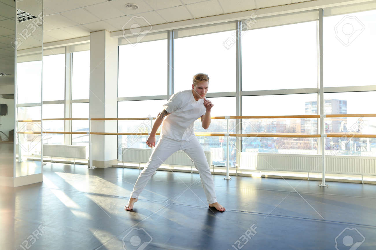 スポーツジムのバレエバレエの近くで体操選手のトレーニング 男は熱心に足のためのストレッチエクササイズを行っています 白いスポーツ ウェアを着たタトゥー集中男性 設備の整ったホール 男性や個々のトレーニングのためのスポーツ服の概念 の写真素材 画像素材