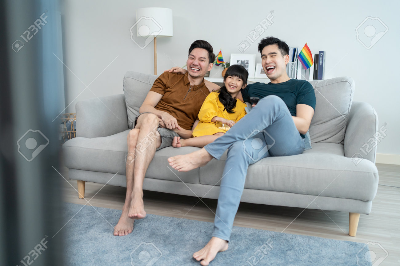 Família do sexo masculino asiático com jovem filha assistir filme na sala de estar. criança menina adorável sentar-se no sofá com atraente casal romântico masculino desfrutar de show de comédia na tv e comer pipoca juntos. - 186124317 Família do sexo masculino asiático com jovem filha assistir filme na sala de estar. criança menina adorável sentar-se no sofá com atraente casal romântico masculino desfrutar de show de comédia na tv e comer pipoca juntos. - 186124317