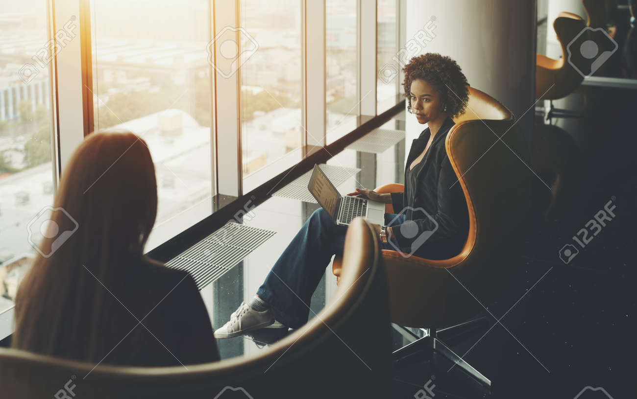 ビジネス会議の 2 つの女性 ノート パソコンと巻き毛と白人と黒人のアメリカ人は 両方高階高層ビルの窓の近く 光が反射してオフィスの高級インテリアのアームチェアに座っています の写真素材 画像素材 Image