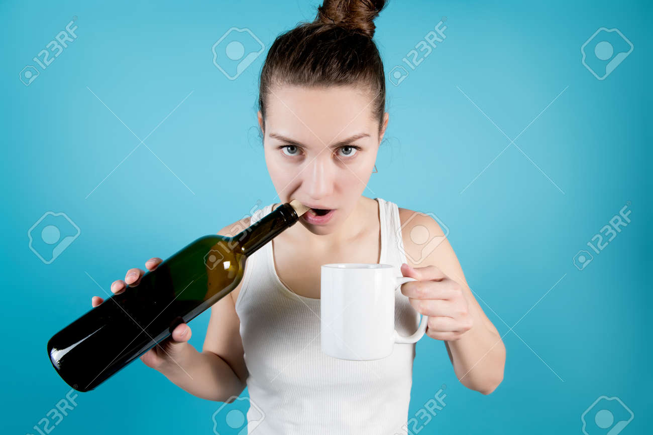 A young woman opens a bottle of wine with her teeth pulling out a cork and is about to pour alcohol into a coffee cup - 150103192 A young woman opens a bottle of wine with her teeth pulling out a cork and is about to pour alcohol into a coffee cup - 150103192
