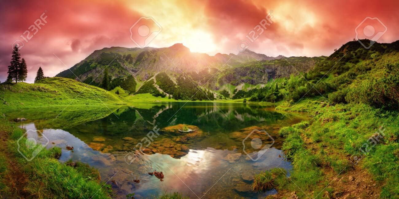 Dramatic sunrise at a lake in the Alps with mountains, red clouds reflected in the clear water and paths leading through the vibrant green grass - 151615450 Dramatic sunrise at a lake in the Alps with mountains, red clouds reflected in the clear water and paths leading through the vibrant green grass - 151615450