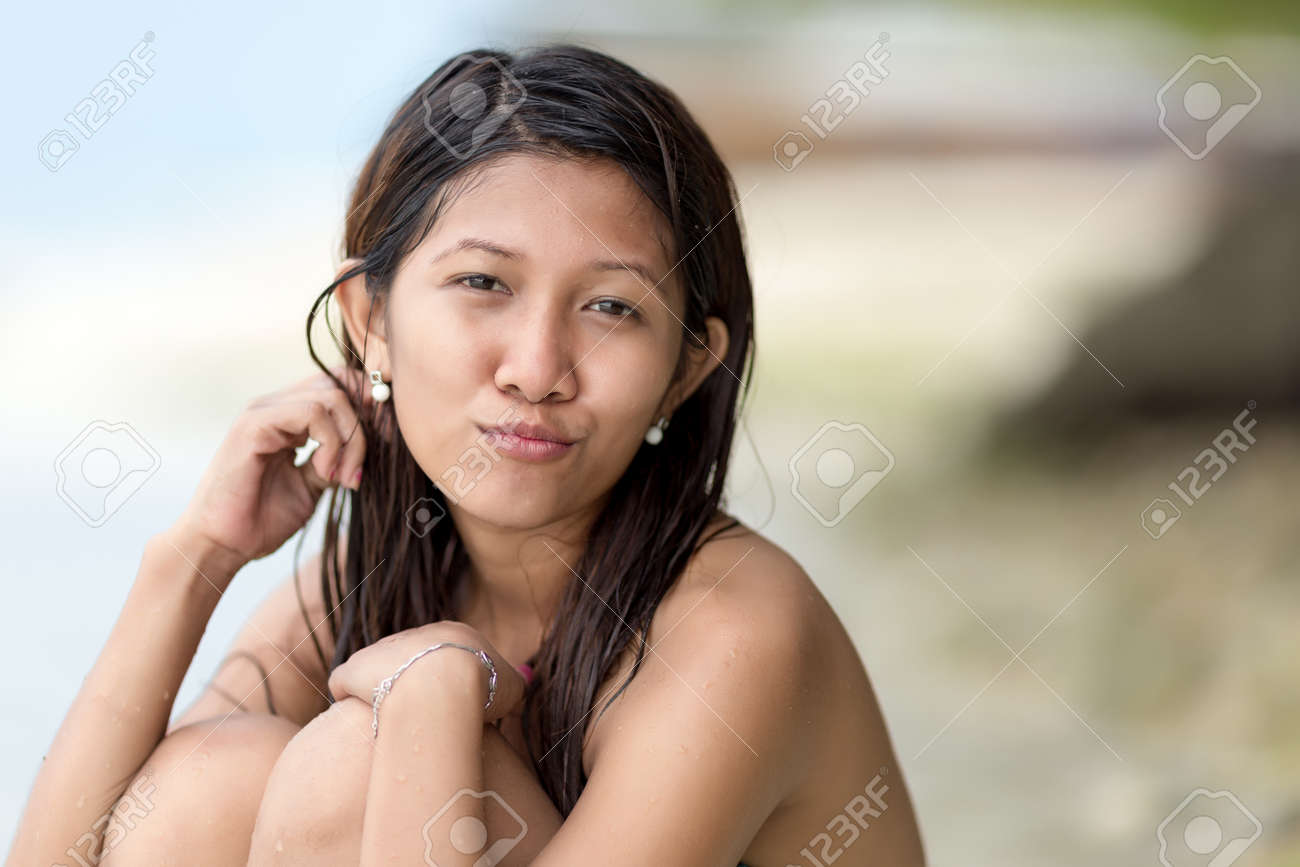 彼女の膝を穏やかな意地の悪い笑みを浮かべてカメラを見てビーチにあごまで曲げて座っている カリスマの美しい若いフィリピン人女性 の写真素材 画像素材 Image