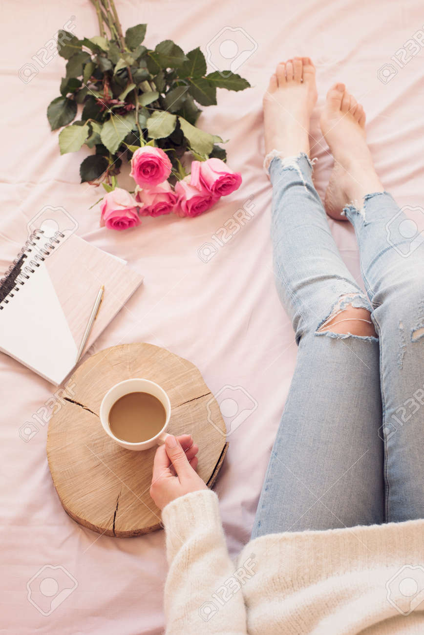 早朝にベッドに入った若い白人女性は 苦しんでいるジーンズと白いセーターを着てコーヒーを飲みます バラとノートの周り 居心地の良いブロガーの朝 の写真素材 画像素材 Image