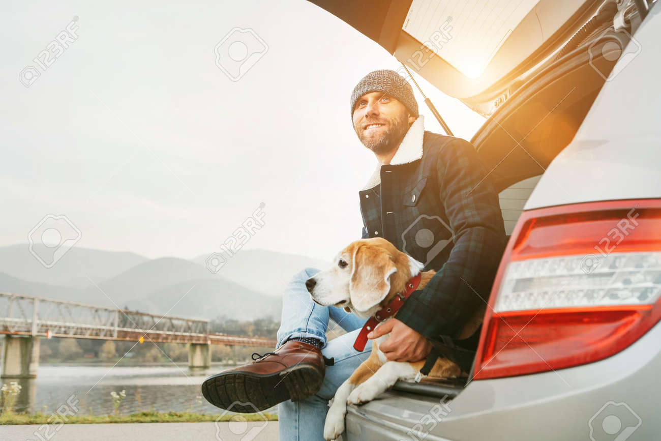 Man with beagle dog siting together in car trunk. Late autumn time - 112309090 Man with beagle dog siting together in car trunk. Late autumn time - 112309090
