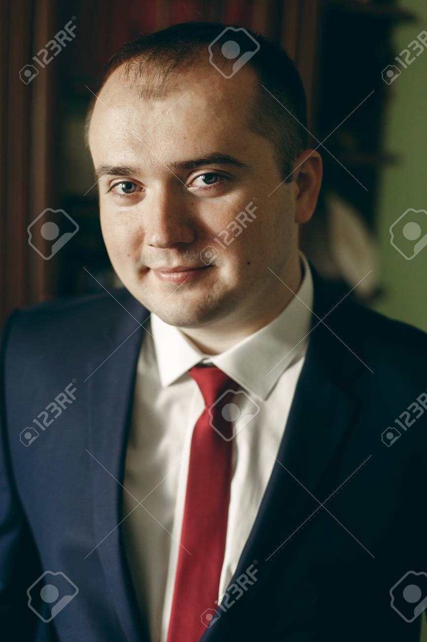 ホテル部屋朝結婚式 幸せなビジネスマンを屋内の肖像画の前に でポーズをとって赤ネクタイ ダークブルーのスーツでスタイリッシュな新郎の幸せ の写真素材 画像素材 Image