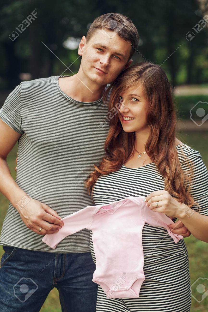 かわいい若い結婚カップル 妊娠中の妻 女の赤ちゃんのための小さなピンクの服を保持している強力な新郎 将来親概念の穏やかな甘い瞬間 の写真素材 画像素材 Image