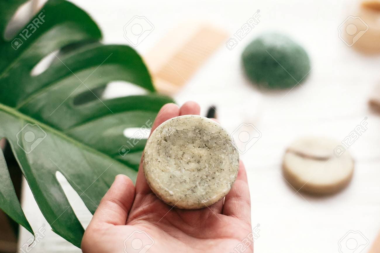 Zero waste. Choice plastic free eco products. Hand holding natural solid shampoo bar on background of bamboo brush, deodorant, sponge on white wood with green monstera leaves. - 111505985 Zero waste. Choice plastic free eco products. Hand holding natural solid shampoo bar on background of bamboo brush, deodorant, sponge on white wood with green monstera leaves. - 111505985