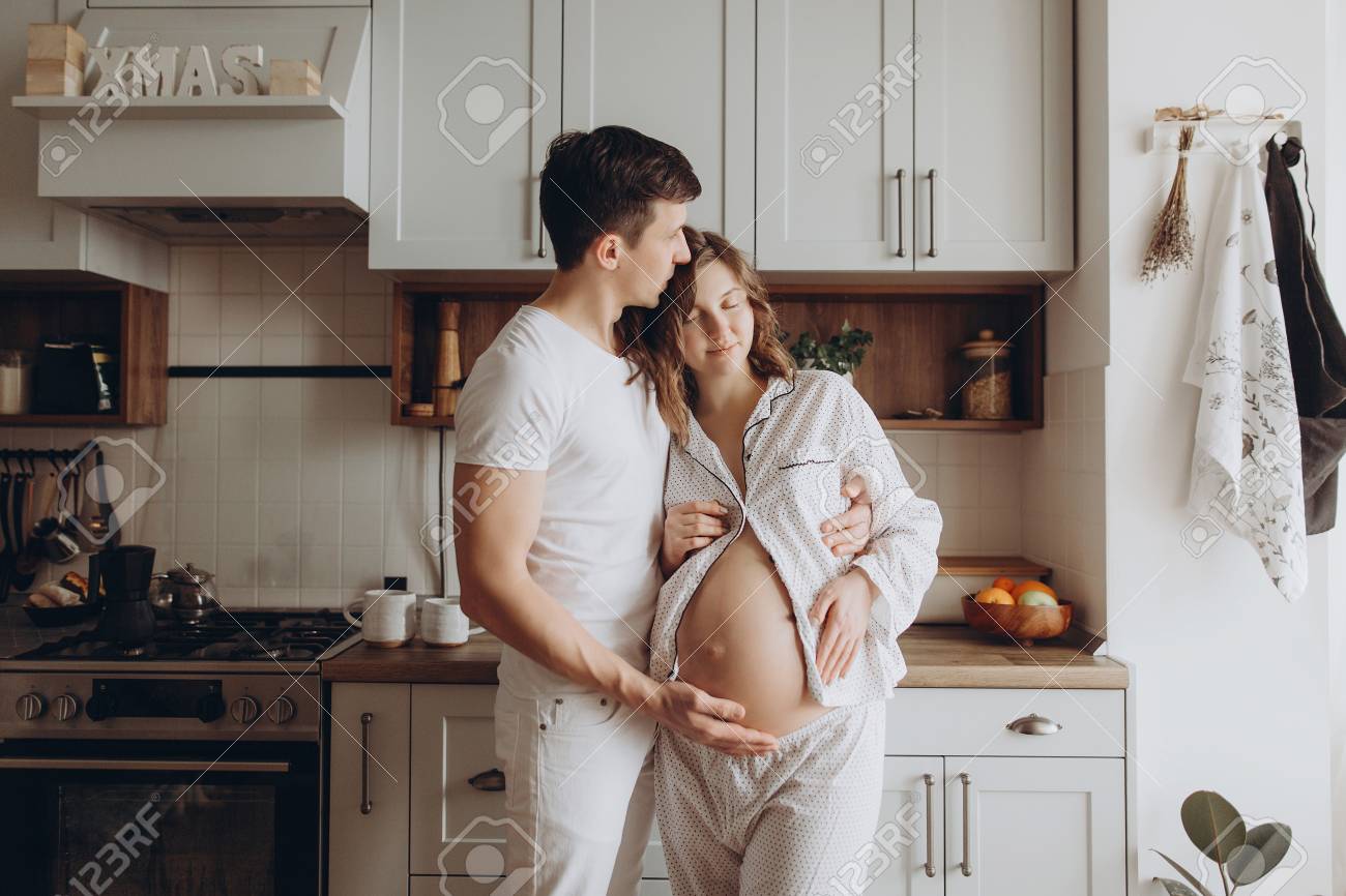 Happy Young Pregnant Couple In White Pajamas Holding Belly Bump