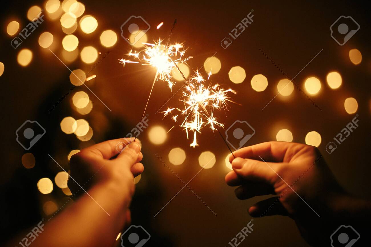 Glowing sparklers in hands on background of golden christmas tree lights, couple celebrating in dark festive room. Happy New Year. Space for text. Fireworks burning in hands. Happy Holidays - 127586424 Glowing sparklers in hands on background of golden christmas tree lights, couple celebrating in dark festive room. Happy New Year. Space for text. Fireworks burning in hands. Happy Holidays - 127586424
