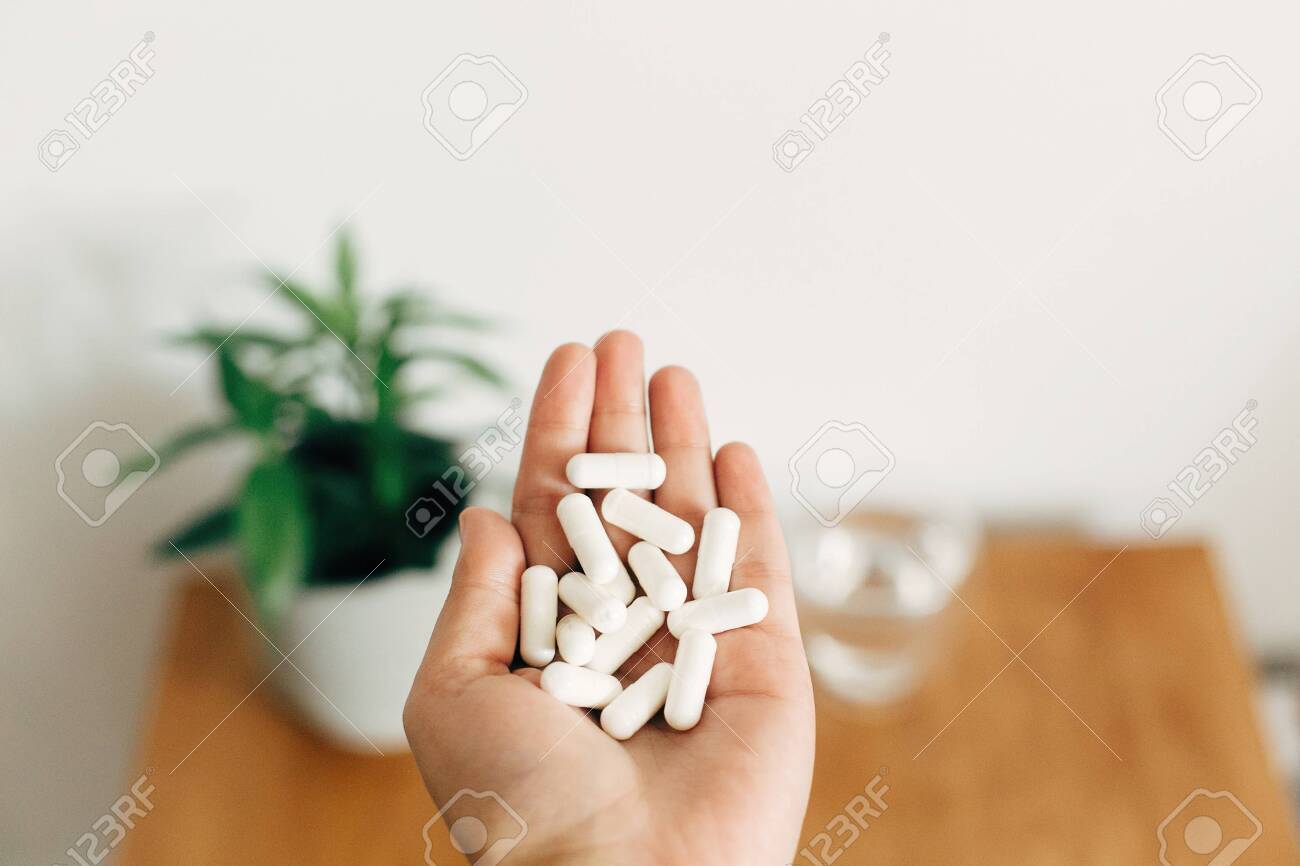 White pills. Dietary supplements.Hand holding magnesium capsules above glass of water on wooden table. Health support and treatment. Biologically active additives - 130447686 White pills. Dietary supplements.Hand holding magnesium capsules above glass of water on wooden table. Health support and treatment. Biologically active additives - 130447686