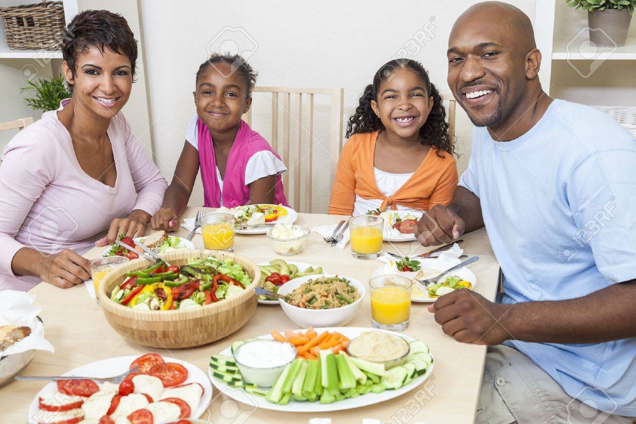An attractive happy African American, smiling family of mother, father, two daughters eating salad and healthy food at a dining table. - 19407100 An attractive happy African American, smiling family of mother, father, two daughters eating salad and healthy food at a dining table. - 19407100