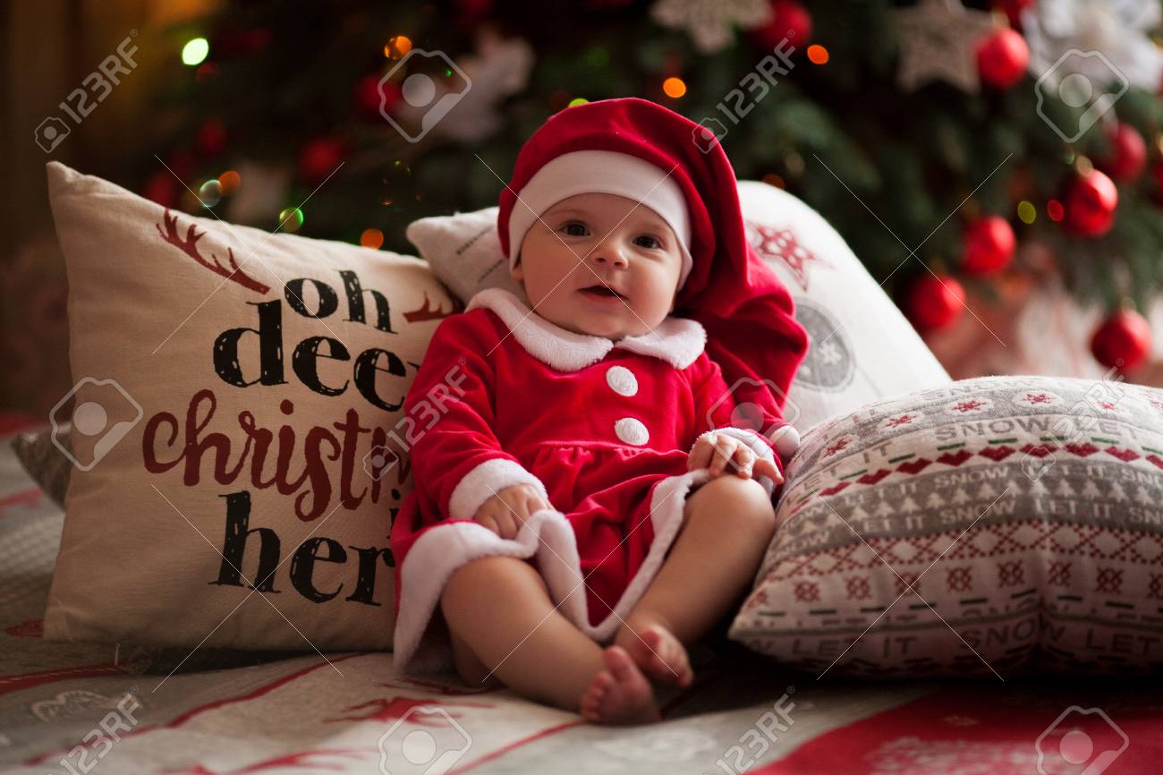 Baby Girl In Santa Claus Costume And Hat Smiles And Sits Barefoot On Bed  Among Pillows Against Background Of Christmas Decorations And Lights. Stock  Photo, Picture And Royalty Free Image. Image 90219998.