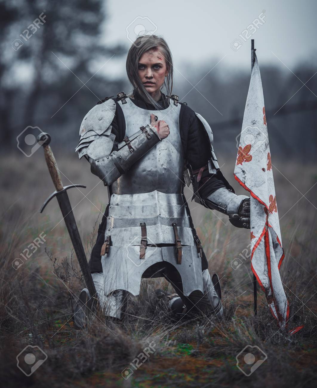 girl in armor kneels with flag in her hands and sword on meadow