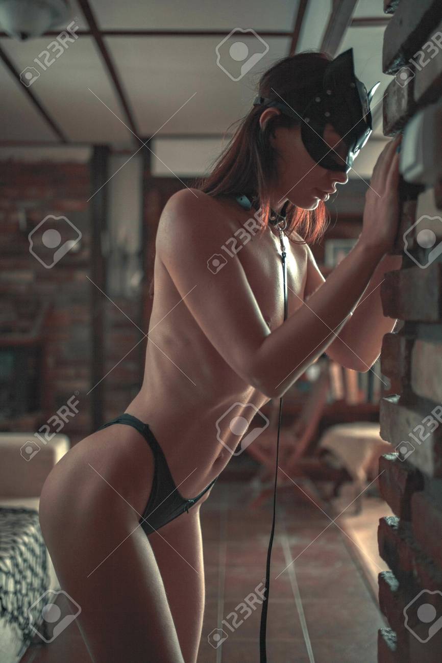 Young Naked Woman In Bikini Stands Near Brick Wall With Black Mask, Collar And Leash On Her Neck. Concept Of Sexual Domination And Bondage