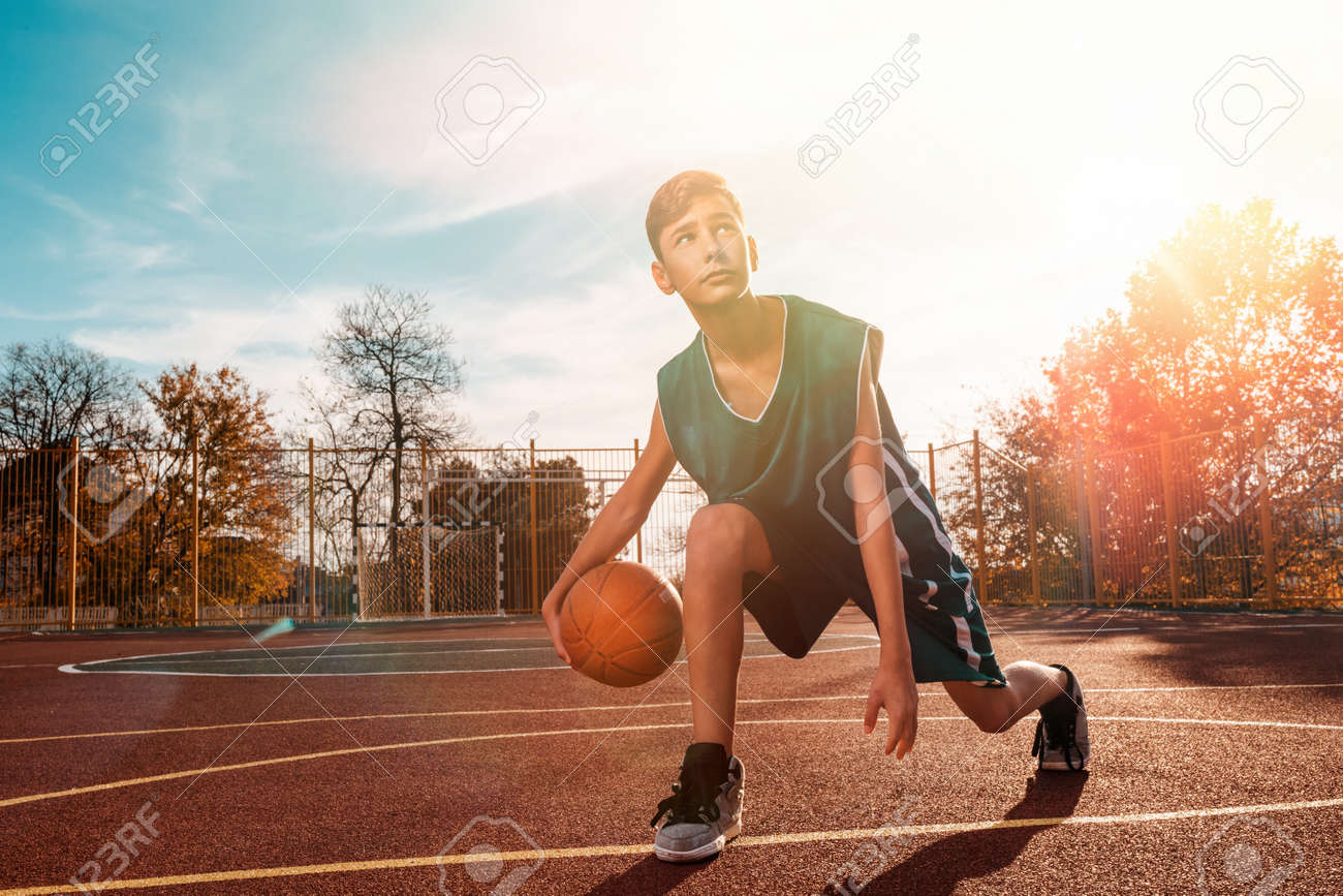 Sports And Basketball. A Young Teenager In A Green Tracksuit Playing  Basketball, Leading The Ball. Blue Sky In The Background And A Sports  Ground In The Background. Sunlight. Copyspace. Stock Photo, Picture, image size:1300x867