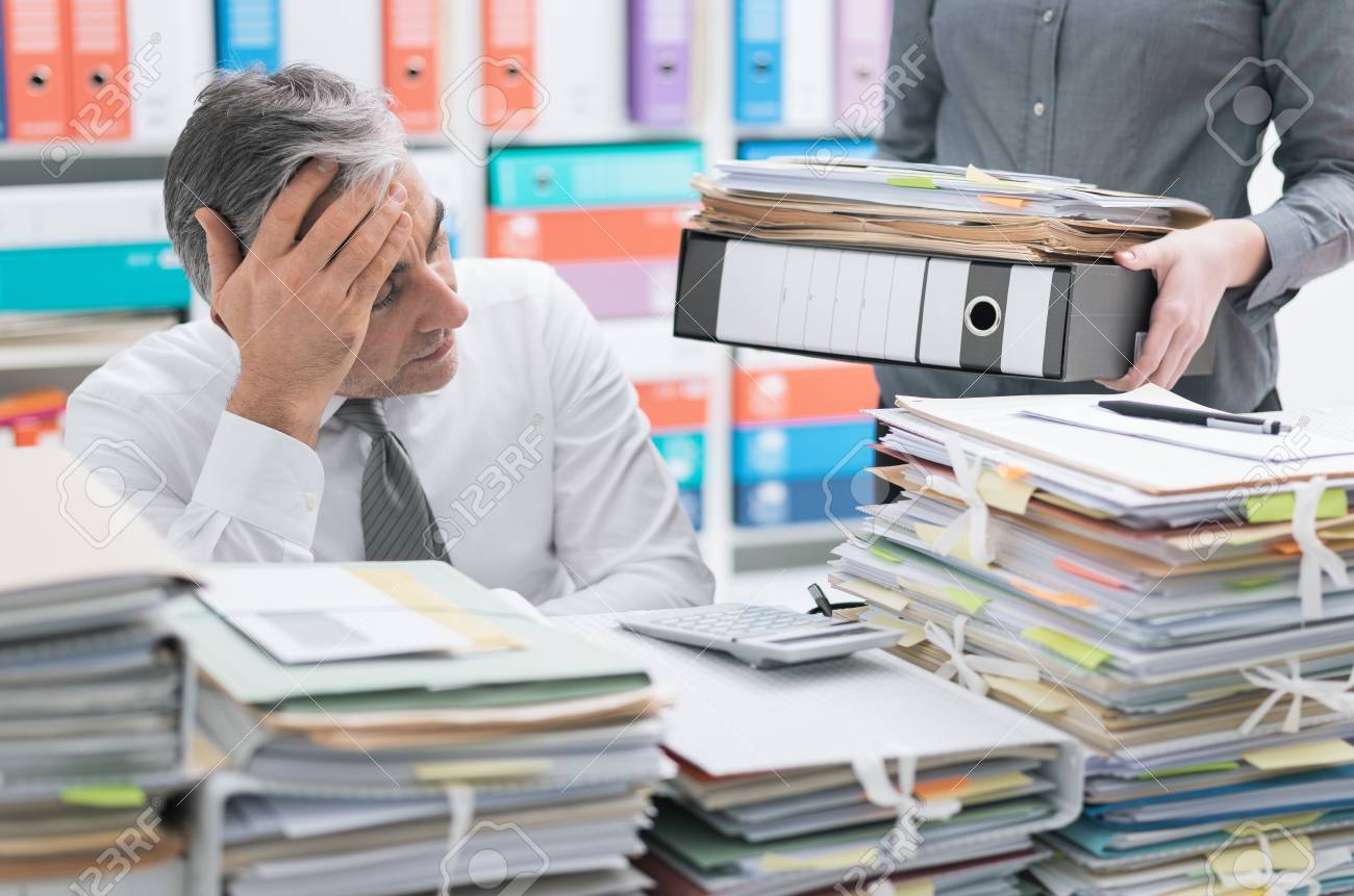 Stressed businessman working at office desk and overloaded with work, the desktop is covered with paperwork, his secretary is bringing more files - 101210305 Stressed businessman working at office desk and overloaded with work, the desktop is covered with paperwork, his secretary is bringing more files - 101210305