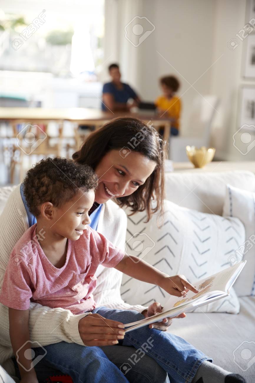 Close up of young mother sitting on a sofa in the living room reading a book with her toddler son, sitting on her knee, father and daughter sitting at a table in the background, vertical - 118101113 Close up of young mother sitting on a sofa in the living room reading a book with her toddler son, sitting on her knee, father and daughter sitting at a table in the background, vertical - 118101113