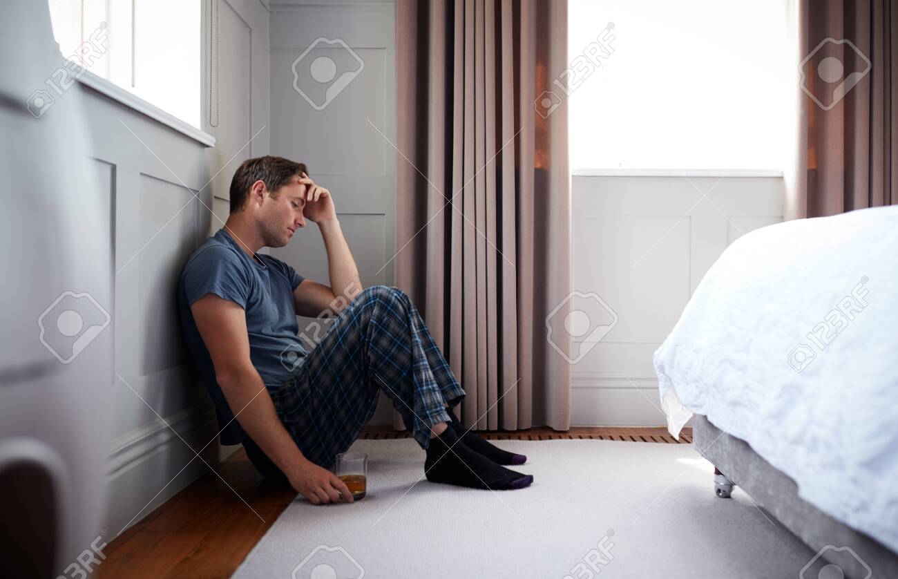 Depressed Man Wearing Pajamas Sitting On Floor Of Bedroom Holding Glass Of Whisky - 129666433 Depressed Man Wearing Pajamas Sitting On Floor Of Bedroom Holding Glass Of Whisky - 129666433