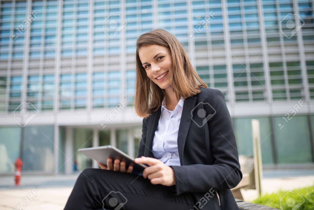 Smiling businesswoman using a digital tablet outdoor sitting on a bench - 154239821 Smiling businesswoman using a digital tablet outdoor sitting on a bench - 154239821