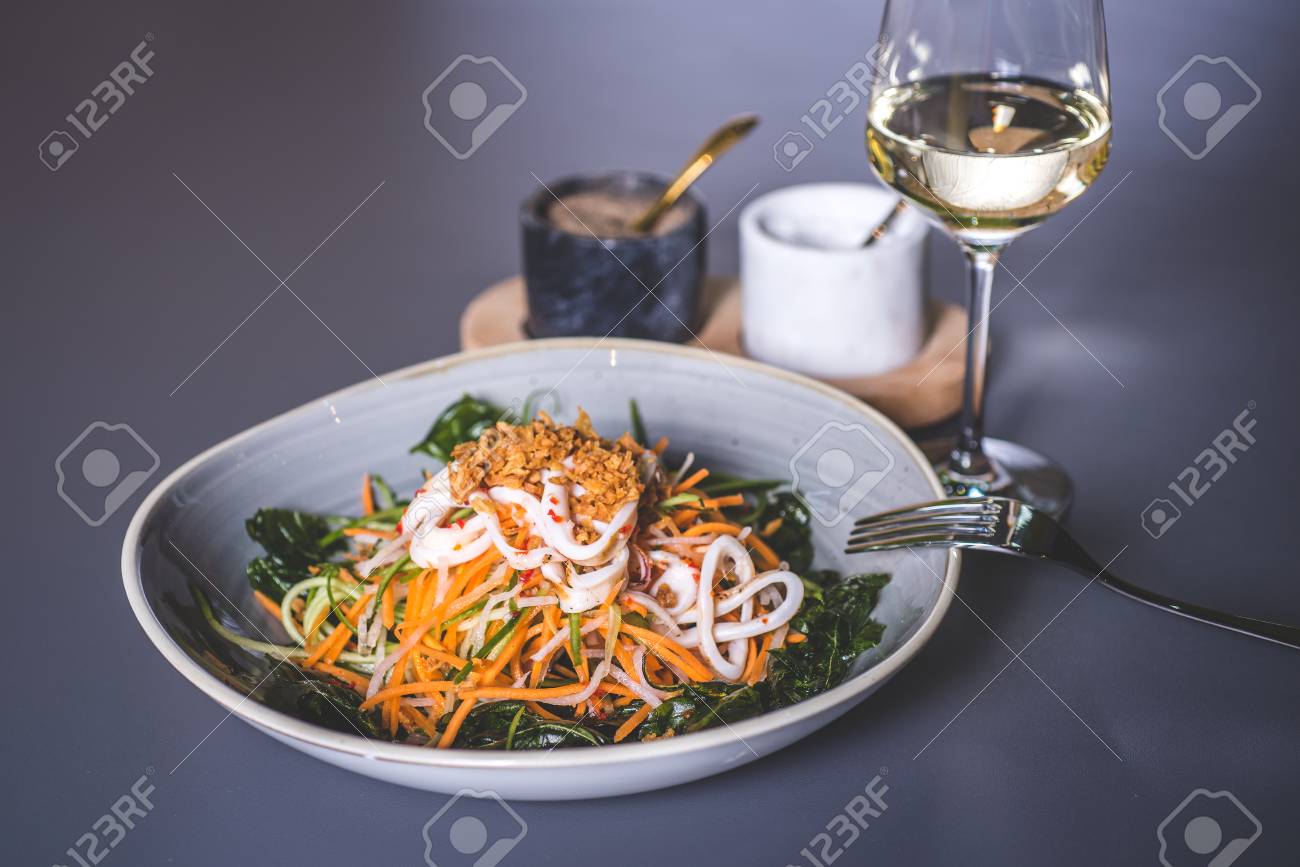 A fork lies on the edge of a plate with a salad. Salad with sea kale and seafood in a ceramic plate. Fougeres with white wine stands on the surface. Marble glasses with spices in the background. - 93005389 A fork lies on the edge of a plate with a salad. Salad with sea kale and seafood in a ceramic plate. Fougeres with white wine stands on the surface. Marble glasses with spices in the background. - 93005389