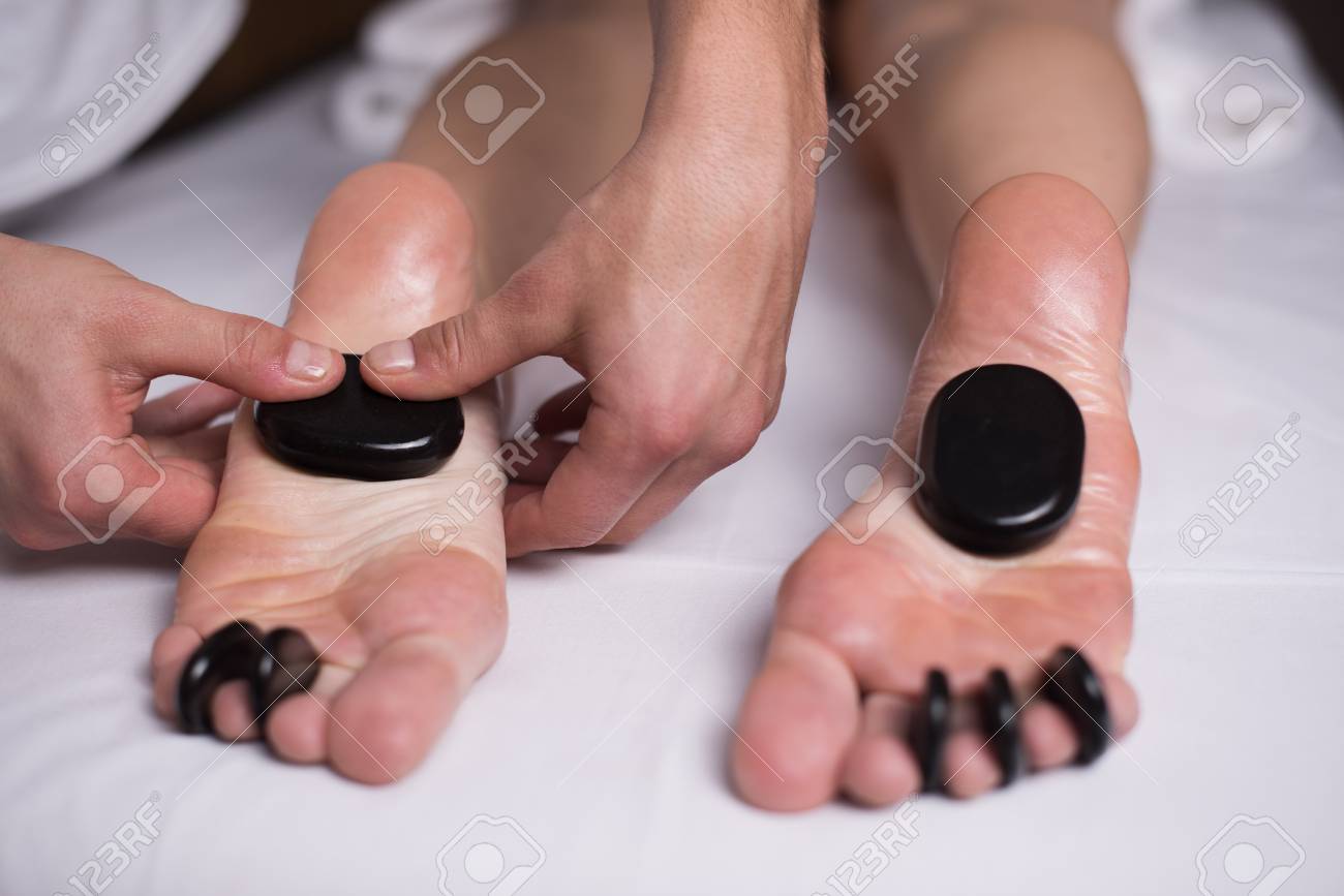 マッサージ師の指は 女性の足に熱い石を押します マッサージ師は熱い石でフットマッサージをします 熱い石は女の子の足とつま先の間に横たわっています の写真素材 画像素材 Image