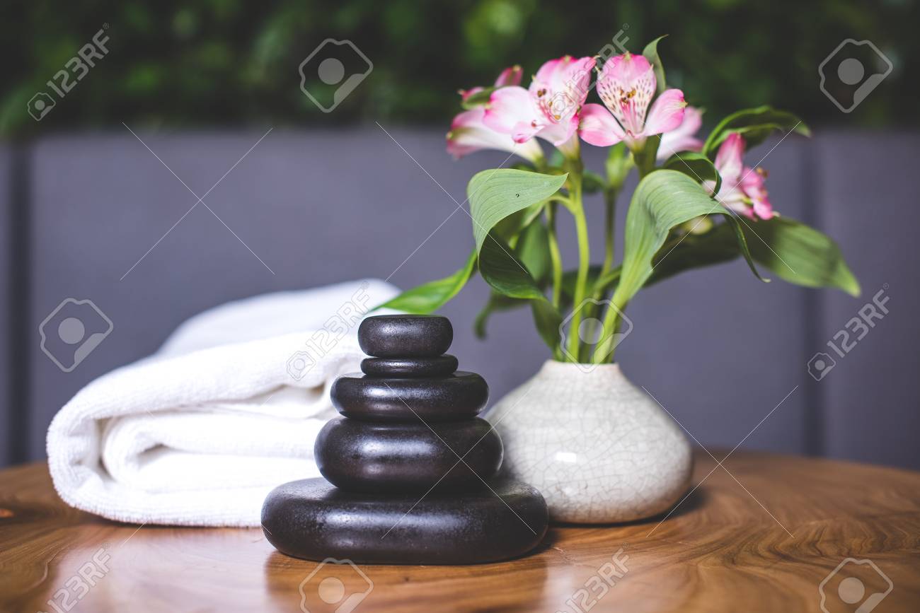 Dark stones for massage are stacked on top of each other on the table. White-pink daffodils stand in a white vase. Pyramids of stones for massage lie on the wooden table. White towels lie in the background. - 94135536 Dark stones for massage are stacked on top of each other on the table. White-pink daffodils stand in a white vase. Pyramids of stones for massage lie on the wooden table. White towels lie in the background. - 94135536