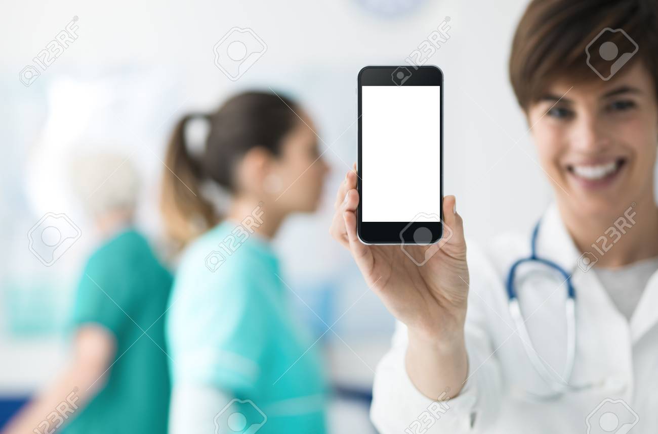 タッチスクリーンスマホや医療スタッフを背景に持つ笑顔の自信満々の女性医師 医療アプリコンセプト の写真素材 画像素材 Image