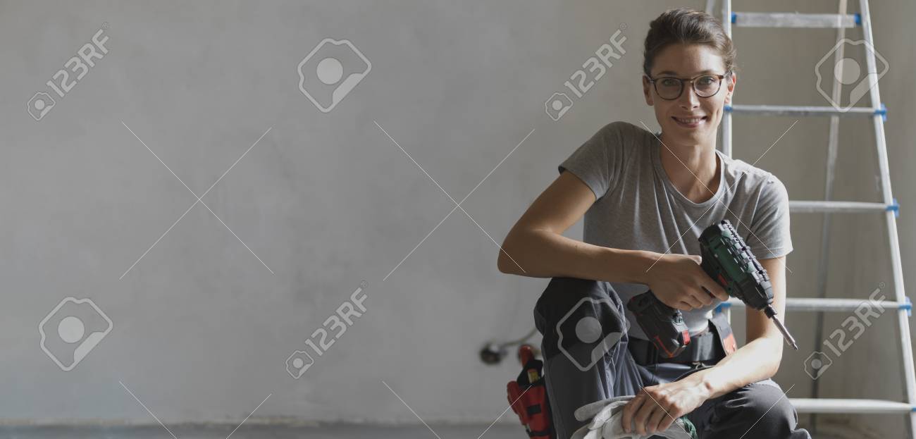 Professional repairwoman with tool belt doing a home renovation, she is posing and holding a drill - 108157682 Professional repairwoman with tool belt doing a home renovation, she is posing and holding a drill - 108157682