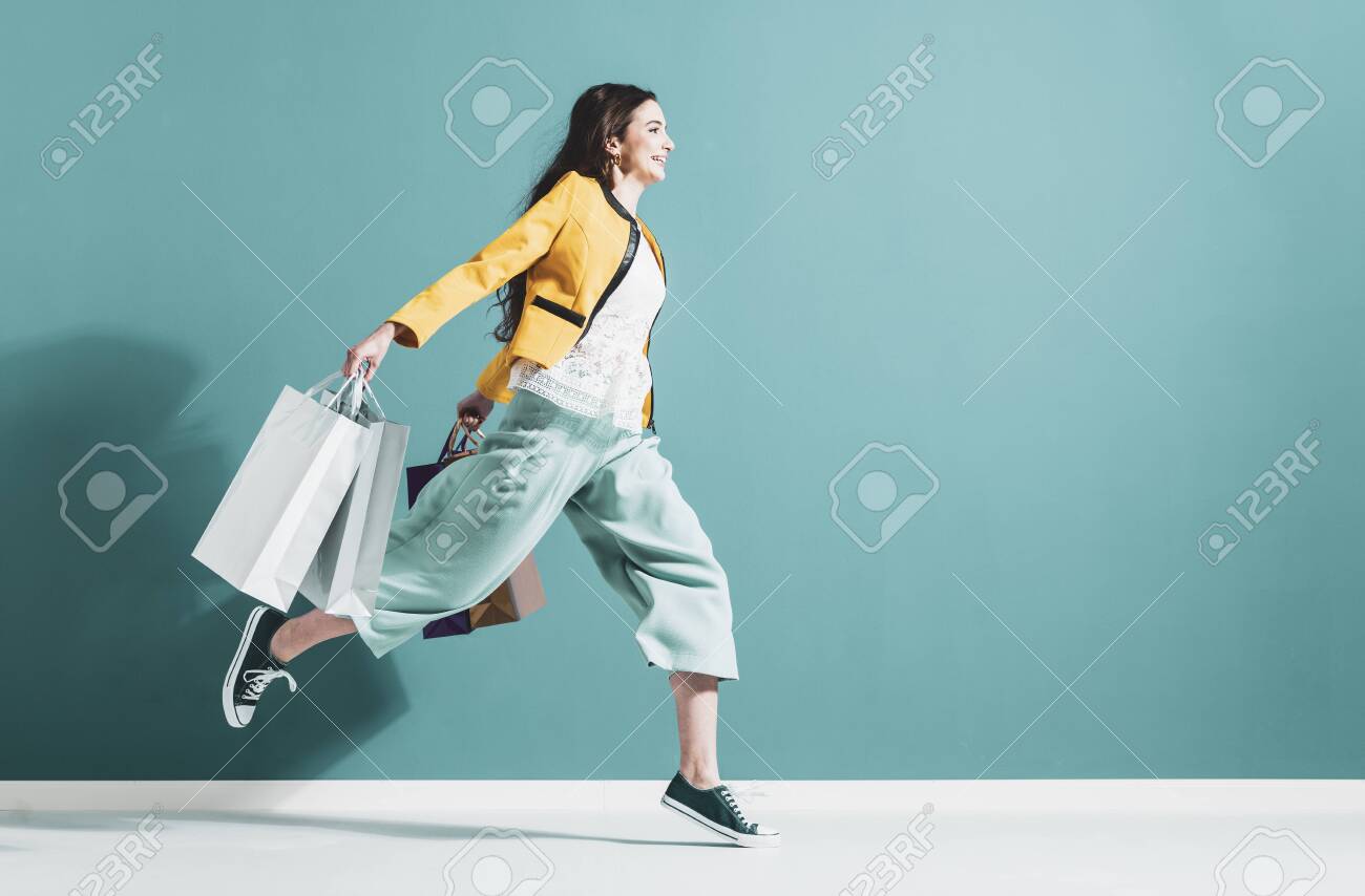 Cheerful happy woman enjoying shopping: she is carrying shopping bags and running to get the latest offers at the shopping center - 125391249 Cheerful happy woman enjoying shopping: she is carrying shopping bags and running to get the latest offers at the shopping center - 125391249