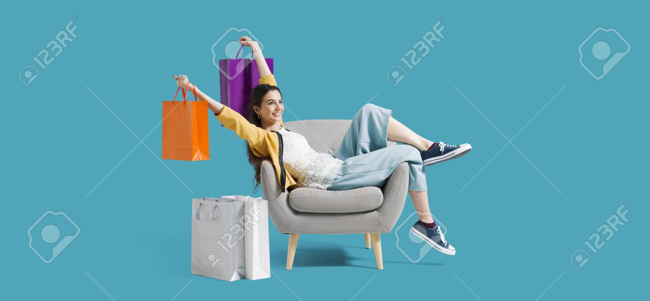 Cheerful happy shopaholic woman with lots of shopping bags, she is sitting on an armchair and celebrating with arms raised - 130088920 Cheerful happy shopaholic woman with lots of shopping bags, she is sitting on an armchair and celebrating with arms raised - 130088920