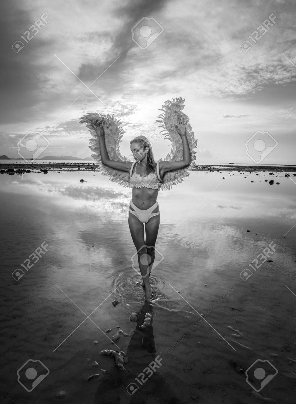 Black and white photo of romantic young beauty as an angel on the beach blonde pretty female woman wearing pink wings feather bra and bikini bottom
