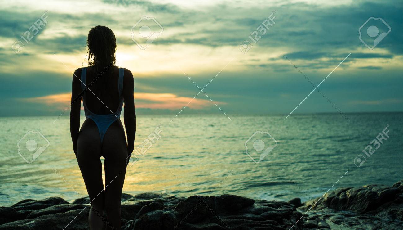 美しい海と夕日を背景とビーチで景色を眺めている濡れた髪と白い水着でセクシーな女性の後ろ姿のシルエット の写真素材 画像素材 Image