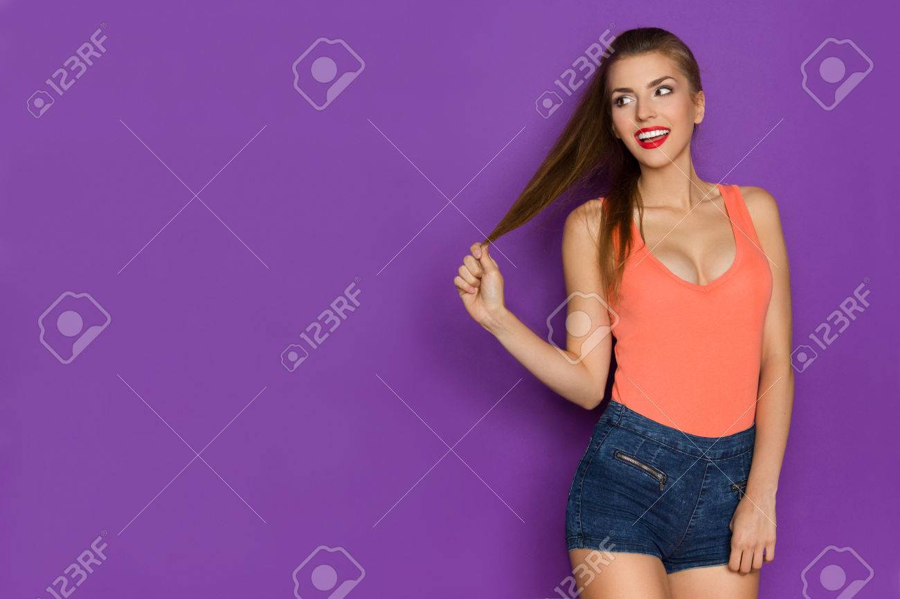 Smiling young woman in orange shirt holding her long brown hair, looking away and talking. Three quarter length studio shot on purple background. - 55320859 Smiling young woman in orange shirt holding her long brown hair, looking away and talking. Three quarter length studio shot on purple background. - 55320859