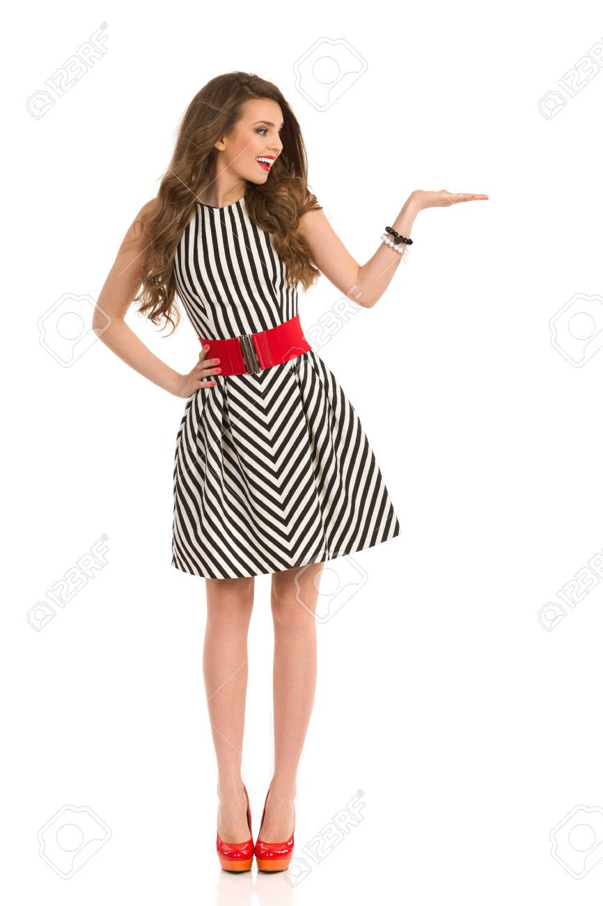 黒と白の縞模様のドレスとハイヒール正面手を上げた 何かを示すと離れて 見てとポーズで魅力的な女性を笑顔します 完全な長さのスタジオ撮影に分離白 の写真素材 画像素材 Image
