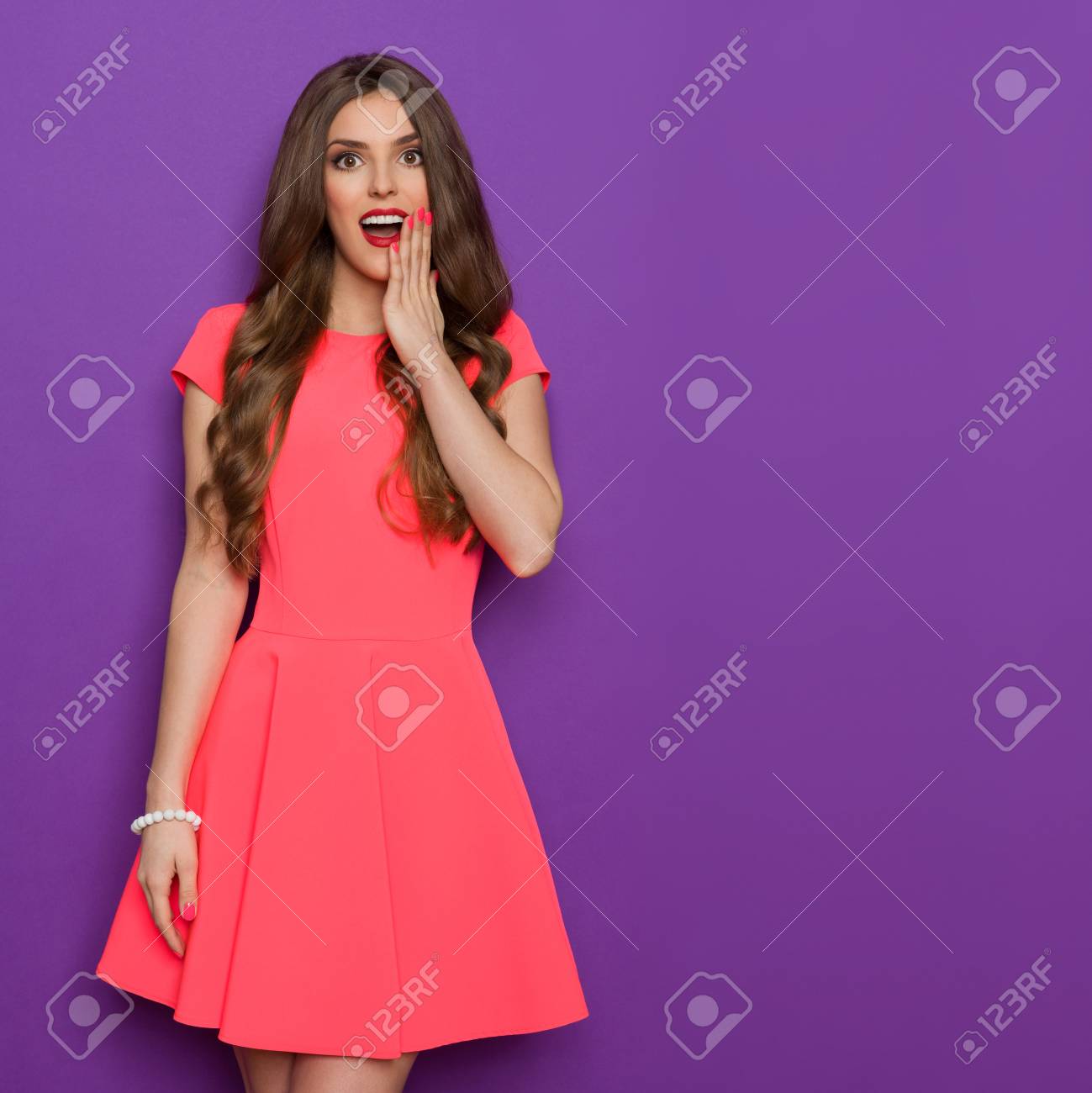 ピンクのミニドレスのあごに手でポーズとカメラ目線で美しい若い女性は驚いた 4 分の 3 の長さスタジオは 紫色の背景に撮影 の写真素材 画像素材 Image