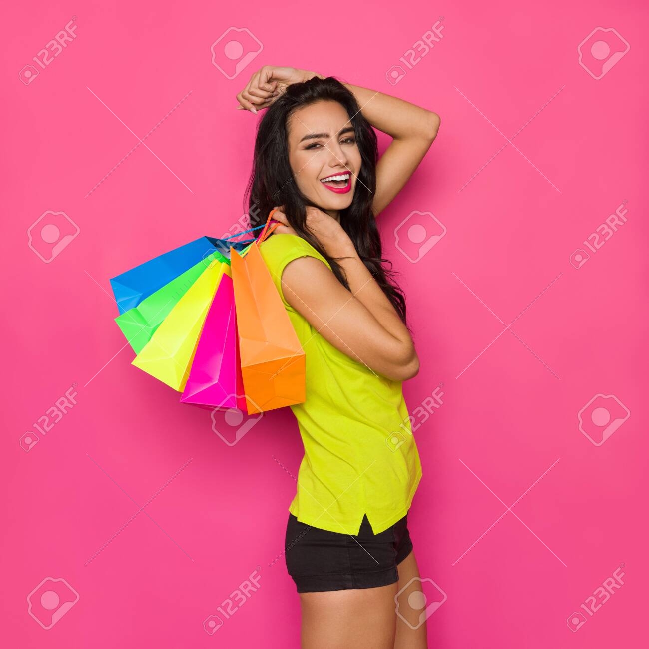 Happy beautiful young woman in vibrant green shirt and black shorts is holding colorful shopping bags on her shoulder and shoutng. Three quarter length studio shot on pink background. - 123523452 Happy beautiful young woman in vibrant green shirt and black shorts is holding colorful shopping bags on her shoulder and shoutng. Three quarter length studio shot on pink background. - 123523452