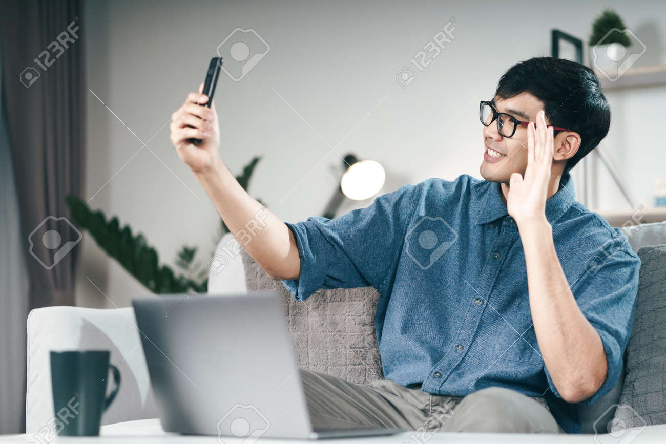 Young Asian man using smartphone for online video conference call and waving hand making hello gesture in the living room. - 171949171 Young Asian man using smartphone for online video conference call and waving hand making hello gesture in the living room. - 171949171