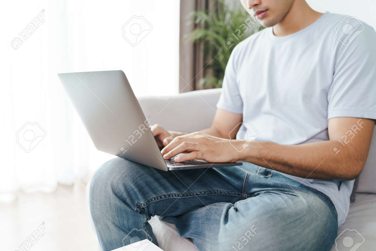 Young man sitting on the sofa at home typing on the laptop keyboard for online working, using internet, freelancer, relaxed, modern job lifestyle, work at home. - 172030051 Young man sitting on the sofa at home typing on the laptop keyboard for online working, using internet, freelancer, relaxed, modern job lifestyle, work at home. - 172030051