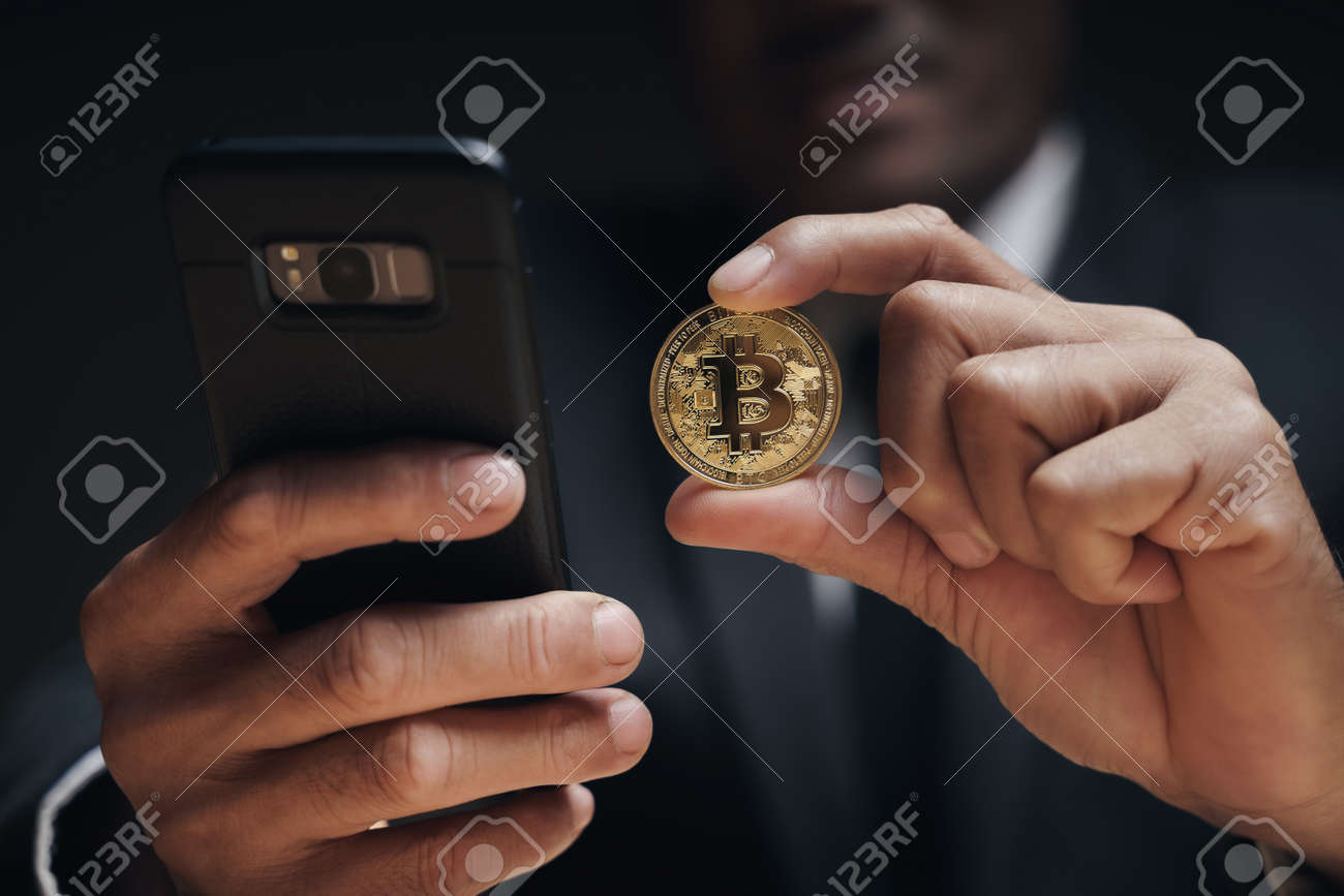Businessman in black suit holding a golden bitcoin using Smartphone with Bitcoin trading chart on the screen to Trading Cryptocurrency, alternative finance and investment Concept. - 172185701 Businessman in black suit holding a golden bitcoin using Smartphone with Bitcoin trading chart on the screen to Trading Cryptocurrency, alternative finance and investment Concept. - 172185701