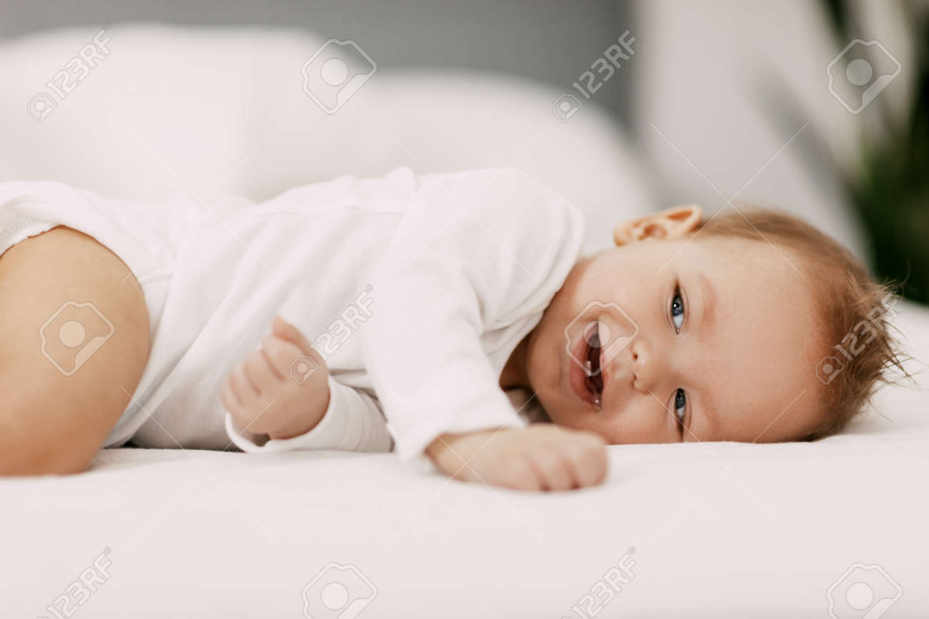 A blue-eyed baby lies in a snow-white bed under a blanket, laughs and indulges - 168313388 A blue-eyed baby lies in a snow-white bed under a blanket, laughs and indulges - 168313388