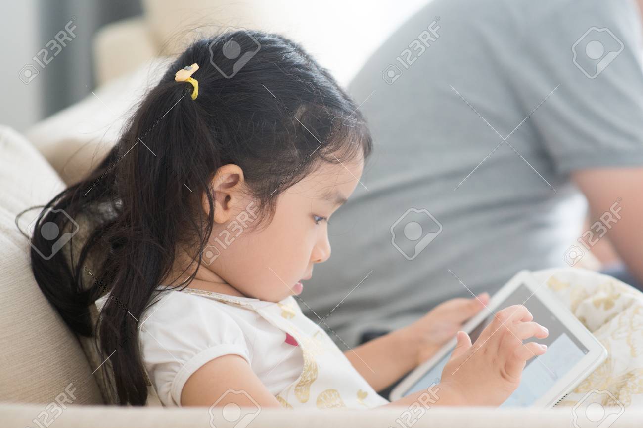 Little girl using digital tablet on sofa. Asian family at home, living lifestyle indoors. - 97097011 Little girl using digital tablet on sofa. Asian family at home, living lifestyle indoors. - 97097011