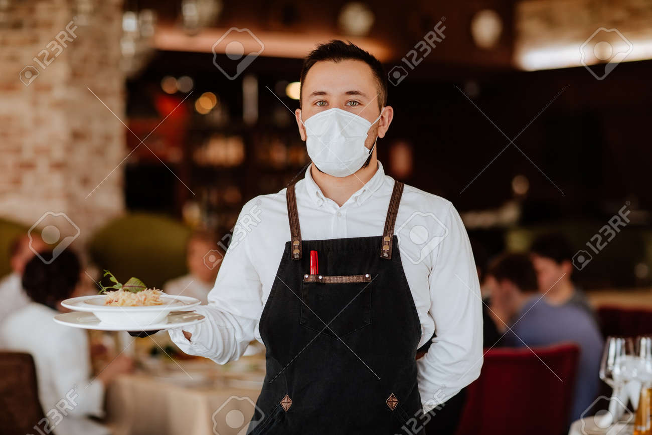 Young handsome waiter in black apron and medical mask holding plate with spaghetti against restaurant background. - 167651307 Young handsome waiter in black apron and medical mask holding plate with spaghetti against restaurant background. - 167651307