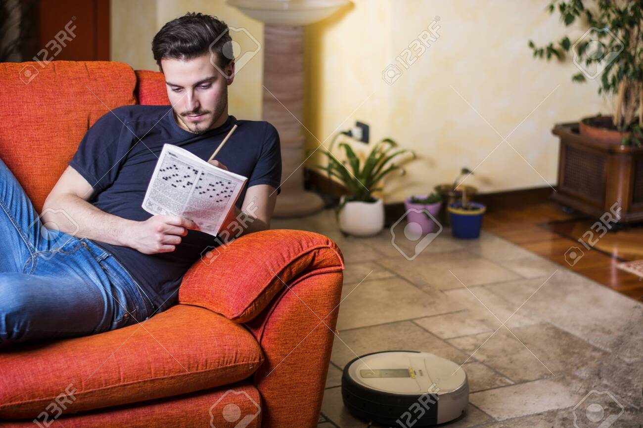 Young Man Sitting Doing A Crossword Puzzle Looking Thoughtfully At A Magazine With His Pencil To His Mouth As He Tries To Think Of The Answer To The Clue Stock Photo Picture